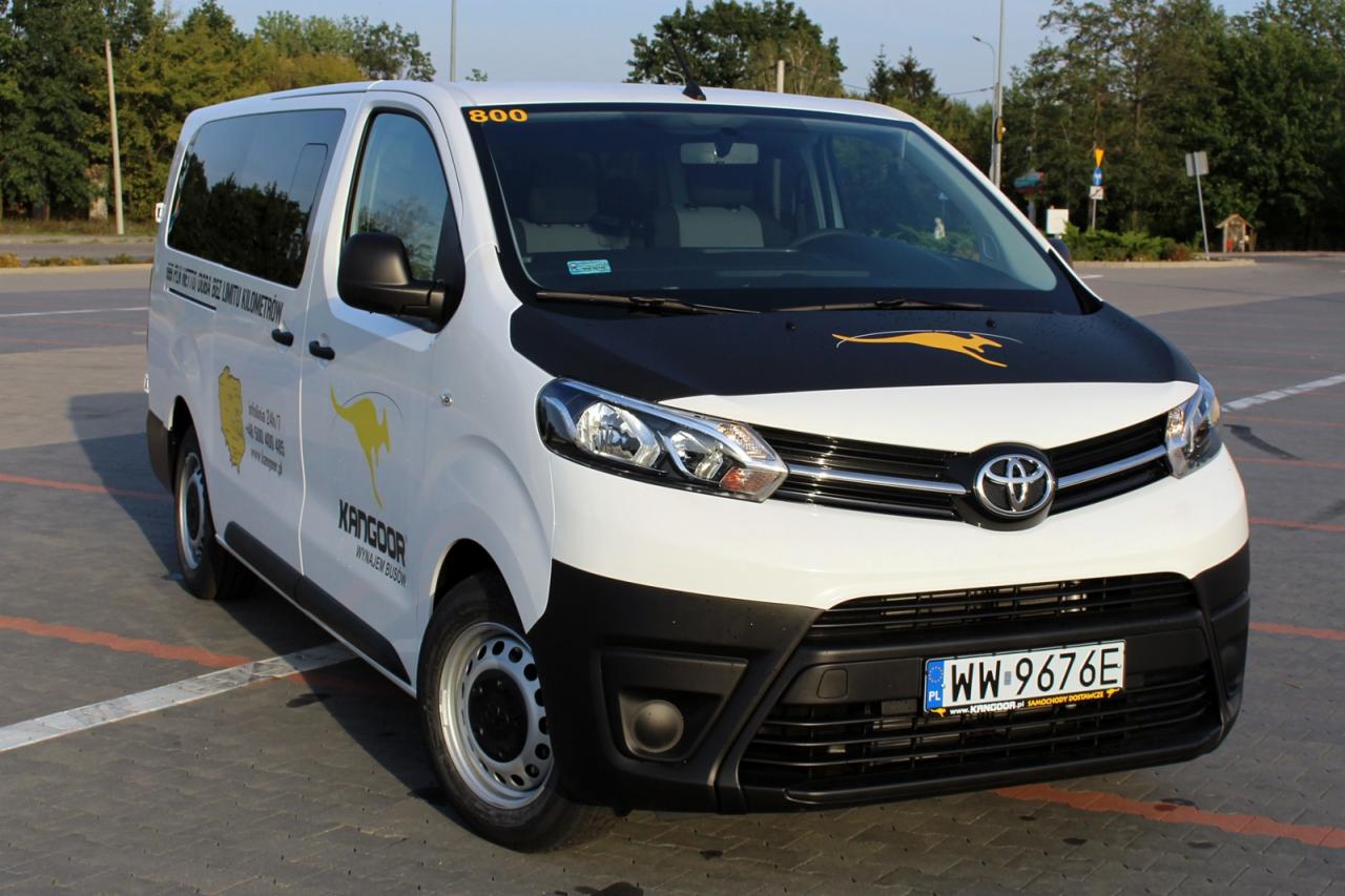 Biały bus marki Toyota z czarnym elementem na masce i żółtym logo kangura, zaparkowany na parkingu.