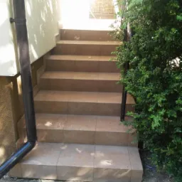 Zewnętrzne schody wejściowe wyłożone jasnobrązowymi płytkami, z czarną rurą spustową po lewej stronie i zielonym krzewem po prawej.