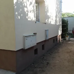 Boczna elewacja budynku po ociepleniu, widoczny fragment okna, skrzynka na listy i małe otwory wentylacyjne w fundamencie, cień drzew na ścianie.