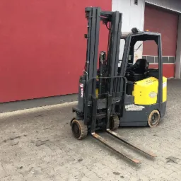 Żółto-szary wózek widłowy elektryczny marki STILL, zaparkowany na kostce brukowej przed czerwono-szarą halą magazynową, widoczne ślady użytkowania na oponach.