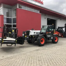 Czarna ładowarka teleskopowa Bobcat z podnośnikiem koszowym na tle budynku z czerwonymi elementami, obok stoją wózki widłowe Linde.