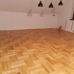 Nowo ułożony parkiet w jodełkę w pokoju na poddaszu z trzema wiszącymi lampami i siedzącym rudym kotem.