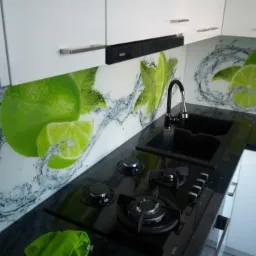 Szklany panel kuchenny z motywem limonki i rozpryskującej się wody, zamontowany nad czarnym blatem z płytą gazową i czarnym zlewem, białe szafki wiszące i stojące.