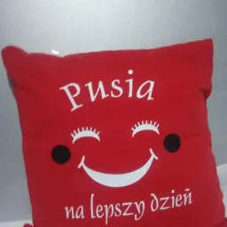 Czerwona poduszka dekoracyjna z białym nadrukiem uśmiechniętej buzi i napisem 'Pusia na lepszy dzień'.