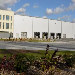 Nowoczesny budynek magazynowy z częścią biurową, widok z zewnątrz, z rampami załadunkowymi i zagospodarowanym terenem zielonym.