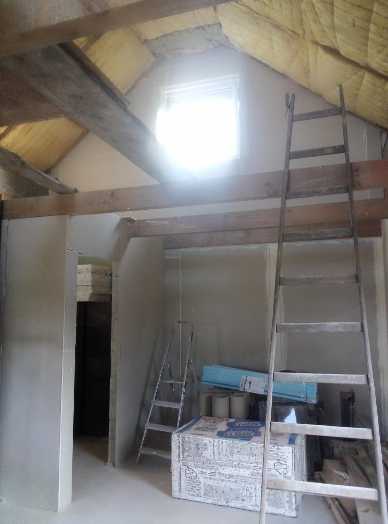 Poddasze w trakcie wykańczania: ocieplenie, płyty gipsowe, drewniane belki, dwie drabiny. Widoczne materiały budowlane i okno w szczycie. Prace remontowe w toku.