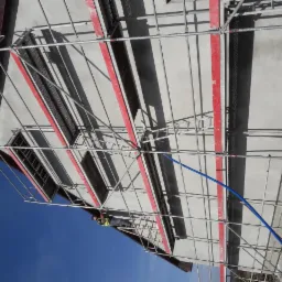 Ściana budynku w trakcie prac wykończeniowych, widoczne rusztowanie z czerwonymi elementami, fragment nieba i pracownik w ubraniu roboczym na rusztowaniu oraz niebieski wąż.