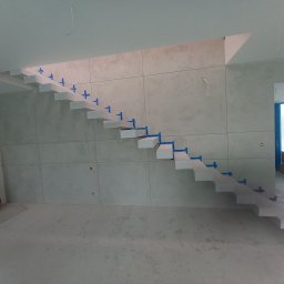 Andrzej Jaworski - Nowoczesne, minimalistyczne schody wspornikowe w trakcie montażu, zabezpieczone niebieską taśmą, na tle ściany wyłożonej płytami w odcieniach szarości.