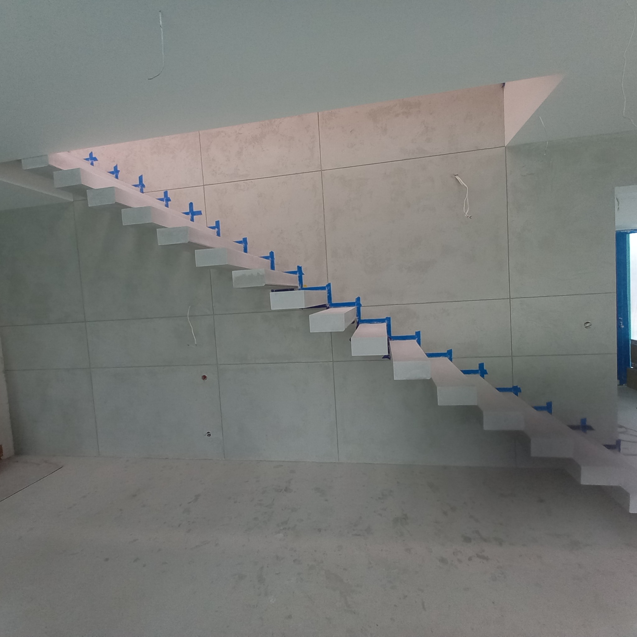 Nowoczesne, minimalistyczne schody wspornikowe w trakcie montażu, zabezpieczone niebieską taśmą, na tle ściany wyłożonej płytami w odcieniach szarości.