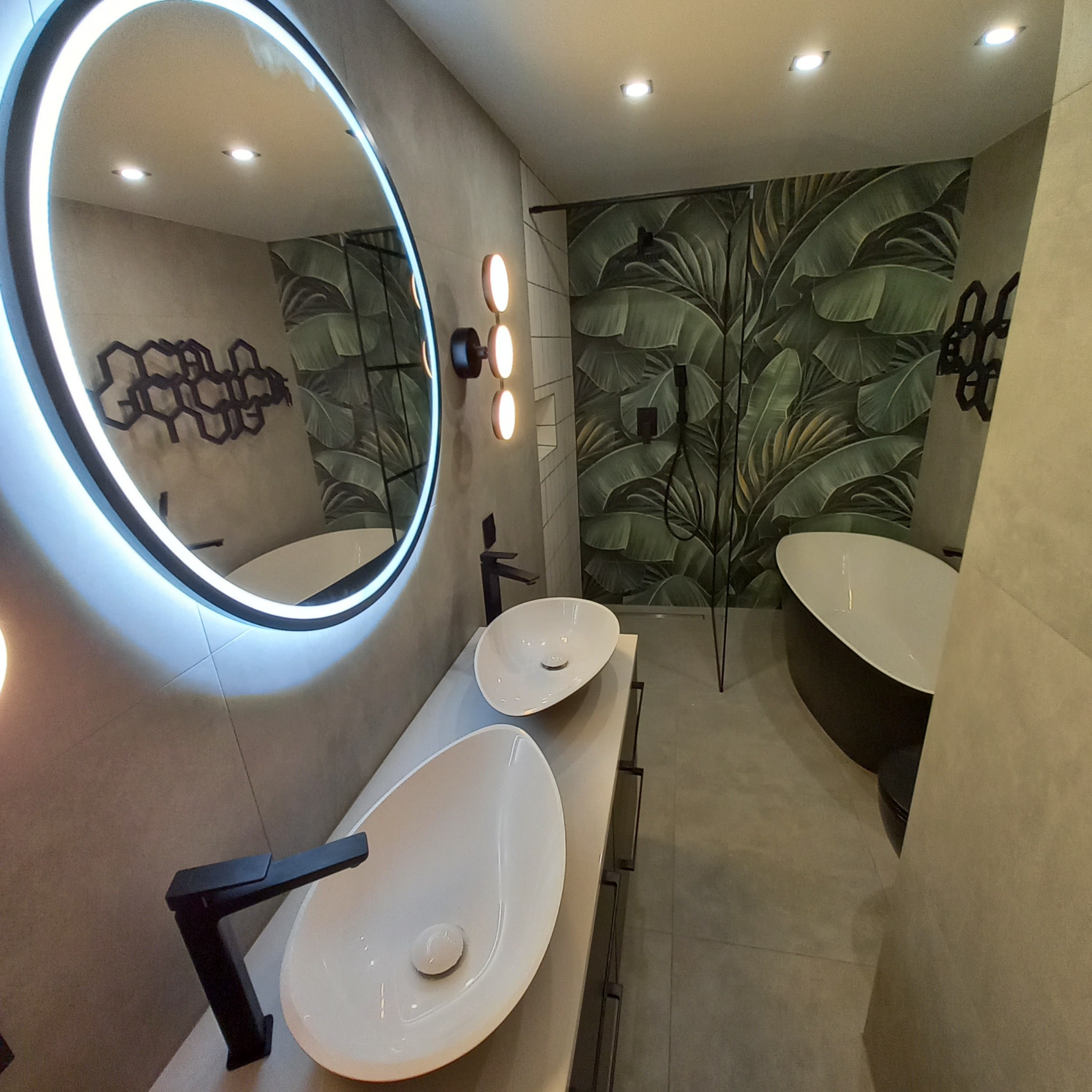 Nowoczesna łazienka z dwoma umywalkami na blacie, okrągłym lustrem z podświetleniem LED i tapetą w motywie liści. Czarna armatura i oświetlenie punktowe.