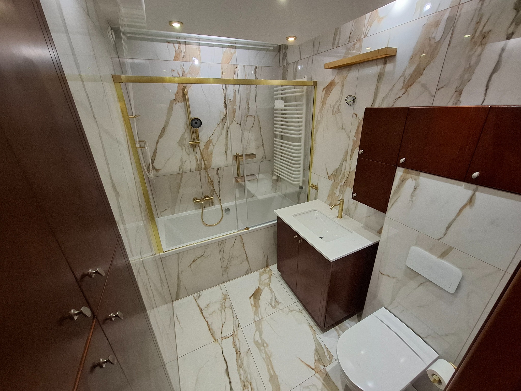 Elegancka łazienka z wanną za szklaną ścianką w złotej ramie, umywalką z szafką i toaletą. Marmurowe płytki na ścianach i podłodze. Ciemne drewniane szafki.