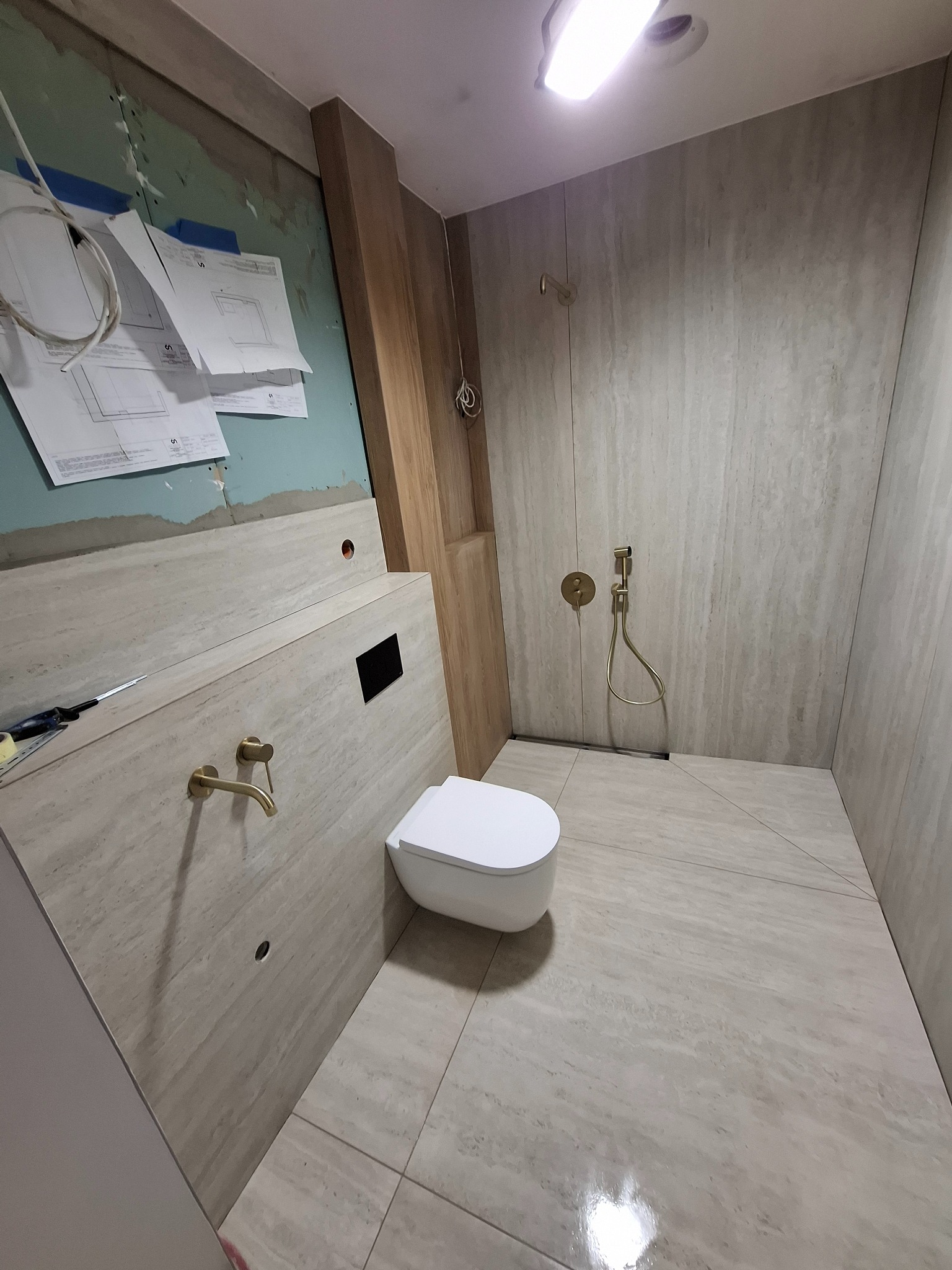 Łazienka w trakcie wykończenia: beżowe płytki imitujące kamień na ścianach i podłodze, wisząca toaleta, złota armatura, widoczne plany i narzędzia na ścianie.