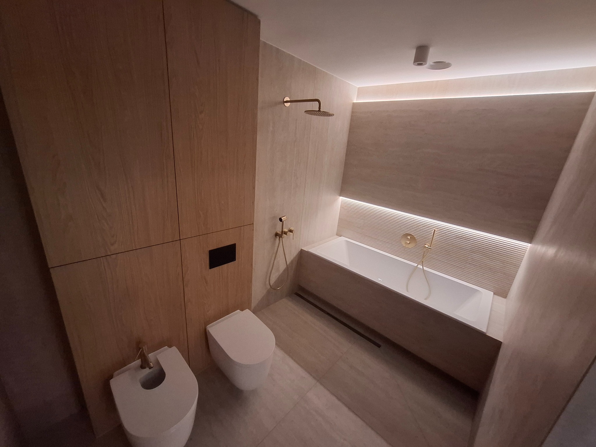 Nowoczesna łazienka z drewnianą zabudową, beżowymi płytkami i złotą armaturą. Minimalistyczny design z podświetleniem LED nad wanną i bidetem.