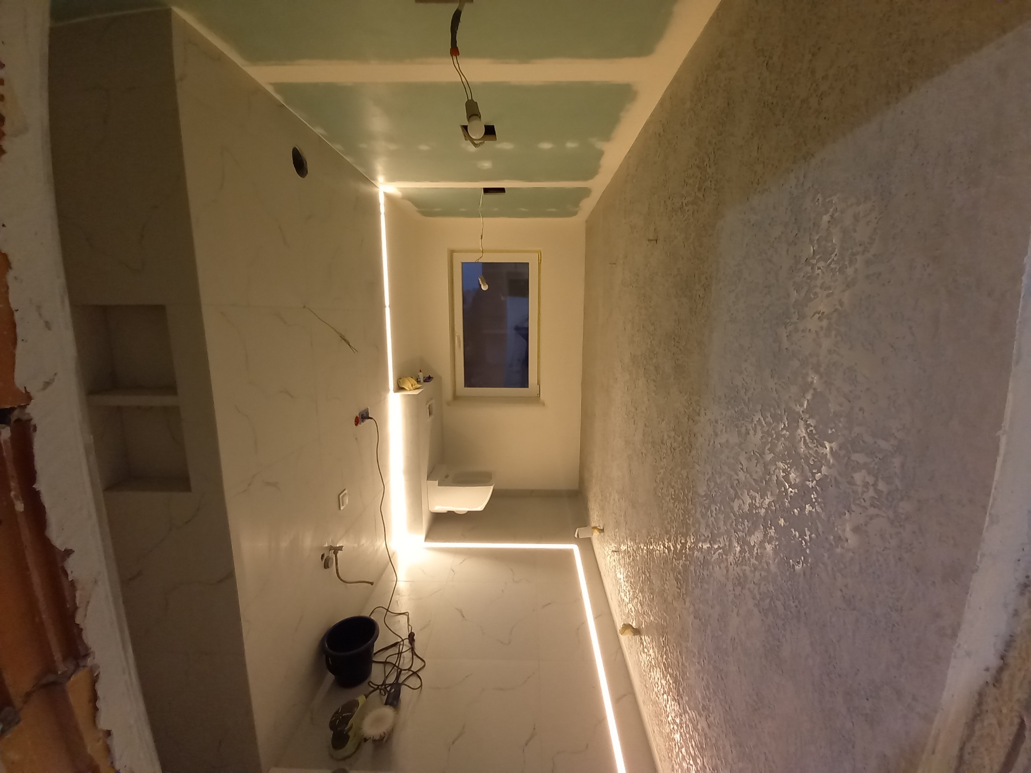 Wnętrze łazienki w trakcie prac wykończeniowych: położone płytki imitujące marmur, ściana z fakturą, podświetlenie LED, sufit z płyt gipsowych, widoczne narzędzia.