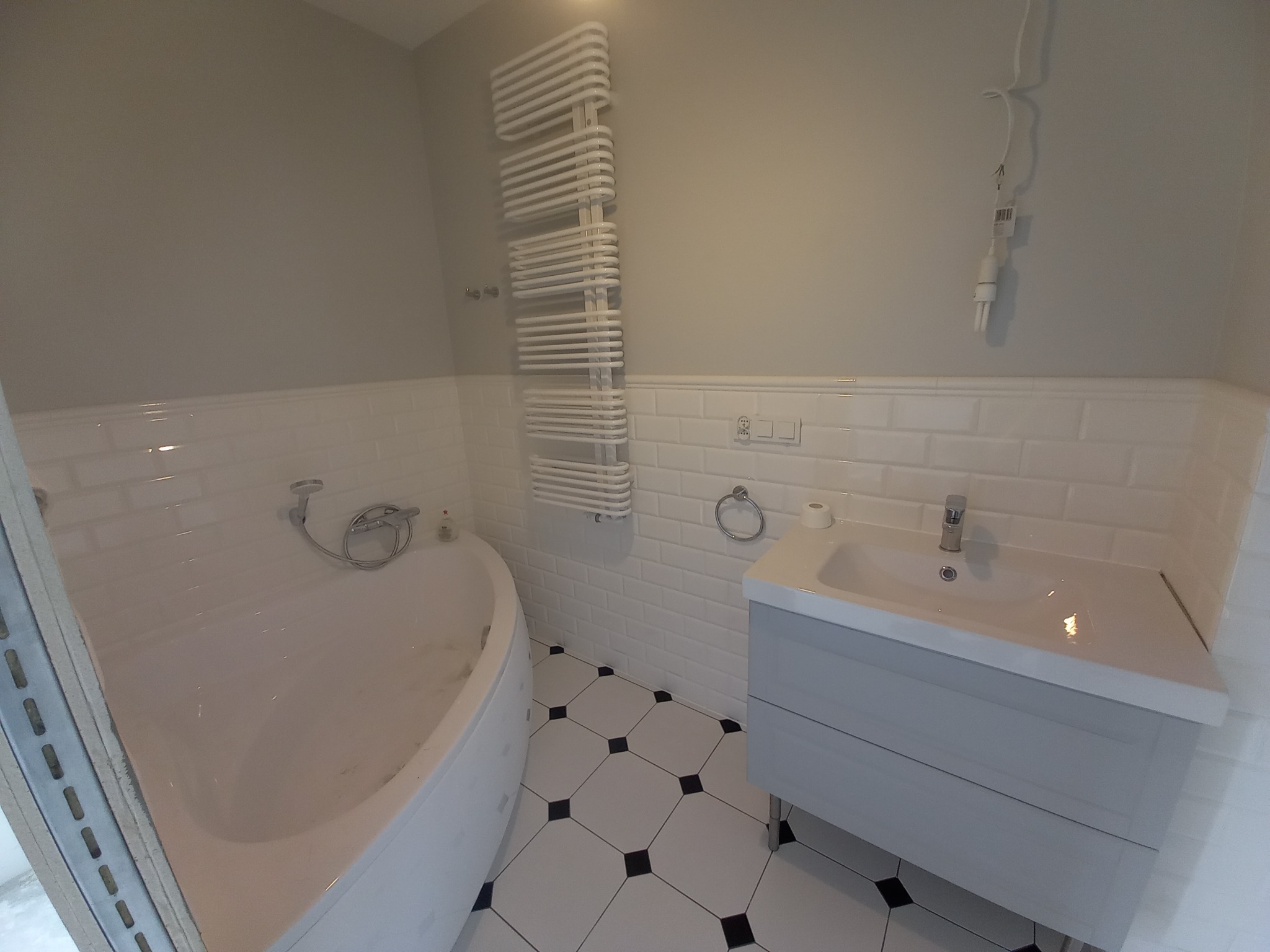 Biała łazienka z wanną, umywalką i grzejnikiem drabinkowym. Ściany wyłożone białymi płytkami imitującymi cegłę, podłoga w czarno-białą szachownicę. Minimalistyczny design.