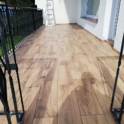 Wykończony balkon w Czarnej Dąbrówce z podłogą wyłożoną płytkami imitującymi drewno, widok zza metalowej balustrady, drabina oparta o ścianę w tle.