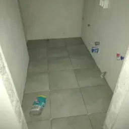 Szare płytki podłogowe ułożone w małej łazience, widoczny worek z klejem do płytek, surowe ściany przygotowane do dalszych prac wykończeniowych.