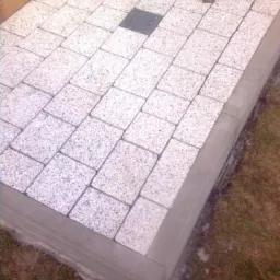 Ułożona nawierzchnia z jasnych kostek brukowych z jedną ciemniejszą kostką pośrodku, obrzeże betonowe, widoczne narzędzia i fragment buta wykonawcy.