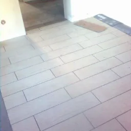 Ułożona podłoga z jasnych, prostokątnych płytek imitujących drewno, widok z góry, w tle przejście do innego pomieszczenia z surową podłogą.