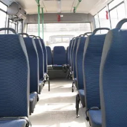 Wnętrze autobusu z rzędami niebieskich, tekstylnych siedzeń z szarymi zagłówkami i czarnymi pasami bezpieczeństwa, widok wzdłuż korytarza w kierunku przedniej szyby.