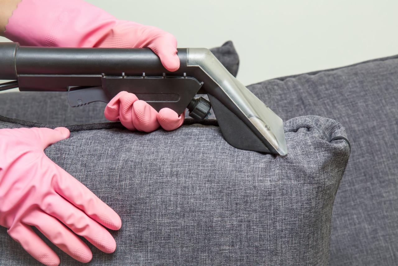 Czyszczenie szarej sofy przy użyciu odkurzacza piorącego, operator w różowych rękawicach ochronnych.