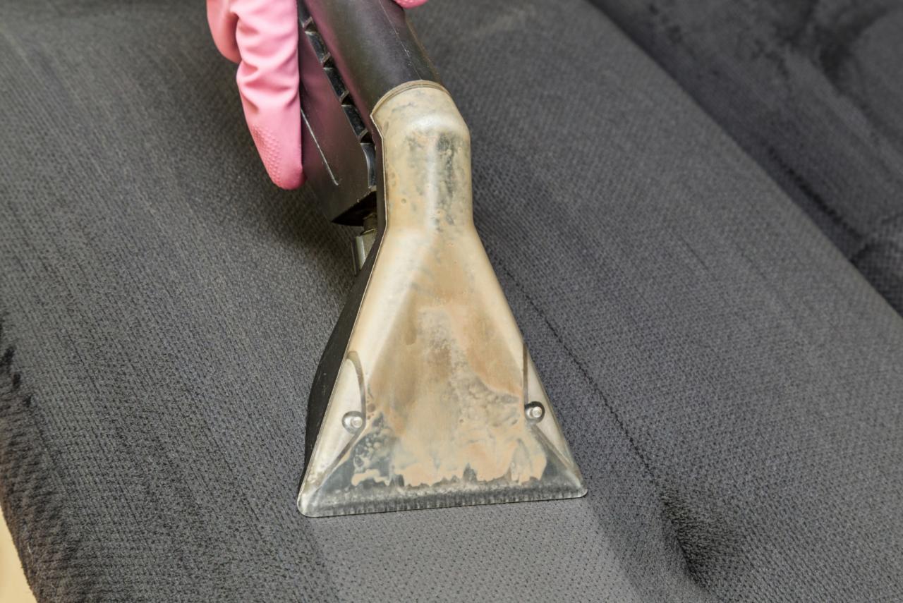 Proces czyszczenia ciemnoszarej kanapy w Gliwicach za pomocą odkurzacza piorącego z widoczną, używaną końcówką i rękawicą ochronną.