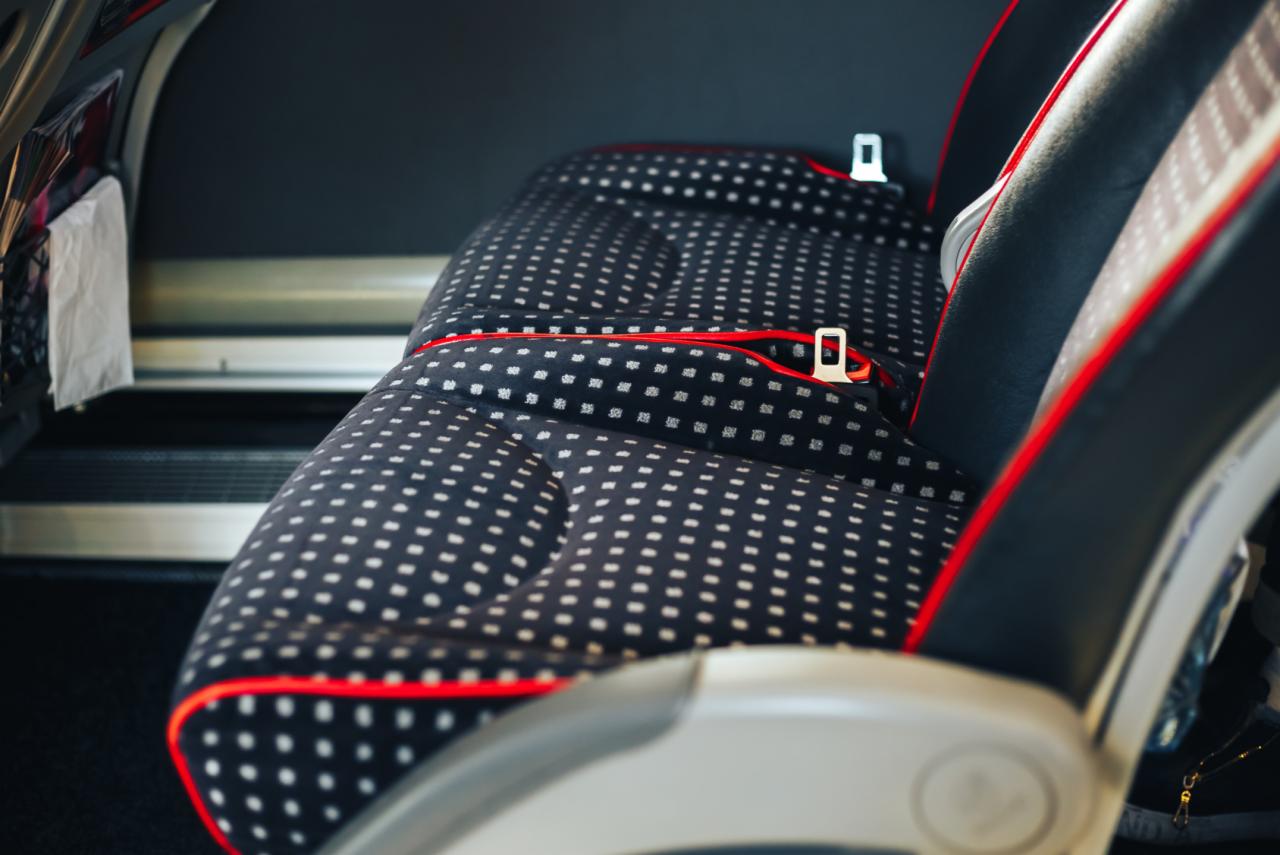 Szczegółowe ujęcie wzorzystego siedzenia autobusu lub pociągu z widocznym pasem bezpieczeństwa i czerwonymi akcentami wykończeniowymi.