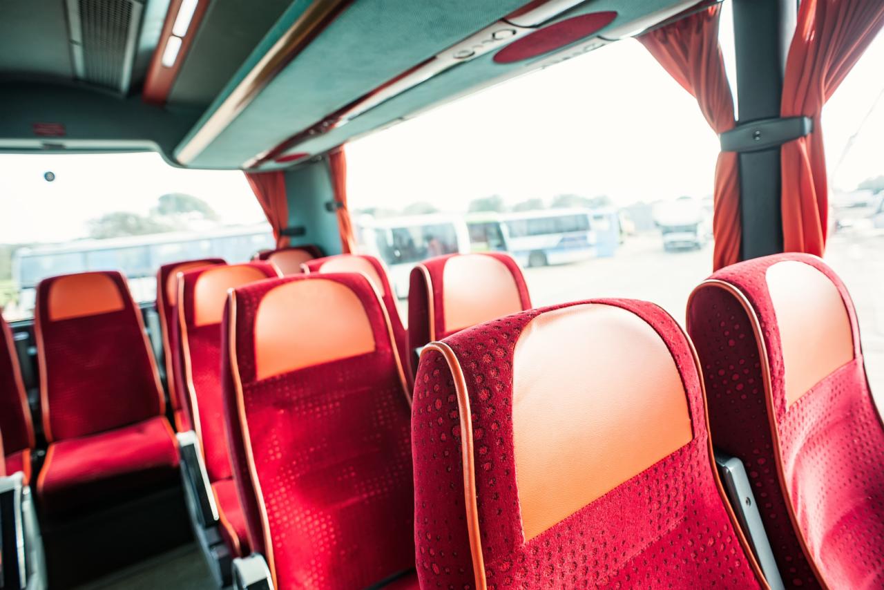 Wnętrze autobusu z rzędami czerwonych foteli z pomarańczowymi zagłówkami, widok na okno i inne autobusy w tle.