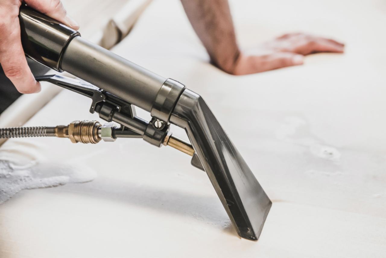 Czyszczenie jasnej tapicerki meblowej parowym odkurzaczem ekstrakcyjnym, widoczna dysza i mokra powierzchnia, dłoń pracownika w tle.