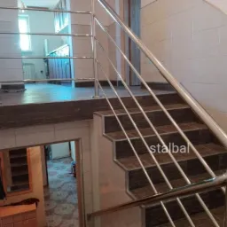 Wewnętrzne schody z brązowymi stopniami i jasnymi płytkami na ścianach, zabezpieczone metalową poręczą z poziomymi prętami, widok z góry schodów na niższy poziom z szafkami i lustrem.