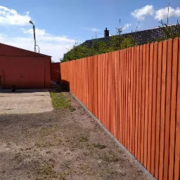 Nowy, pomarańczowy płot z desek sztachetowych, oddzielający posesję z garażem blaszanym, widoczny z perspektywy ziemi w słoneczny dzień.