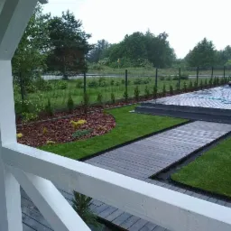 Nowoczesny ogród z drewnianym tarasem i ścieżką, świeżo posadzona trawa, rabata z korą i niskimi iglakami, widok z zadaszonego tarasu z białą balustradą