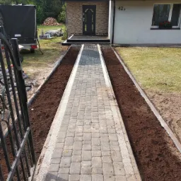 Nowo wyłożona kostka brukowa tworząca ścieżkę prowadzącą do domu z numerem 422, z przygotowanymi rabatami po bokach, widok zza metalowej bramy.