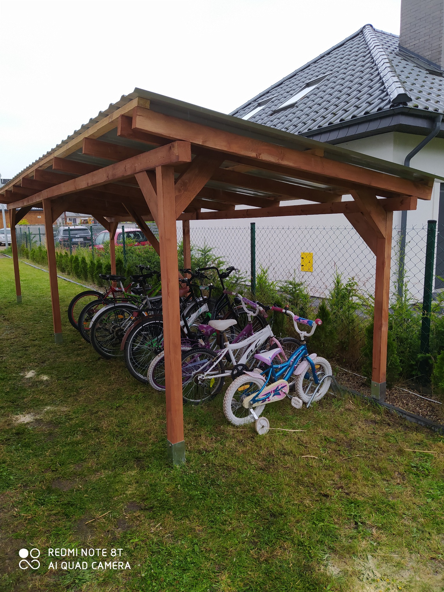 Zadaszona wiata rowerowa z drewnianą konstrukcją i metalowym dachem, stojąca na trawniku przed domem jednorodzinnym.