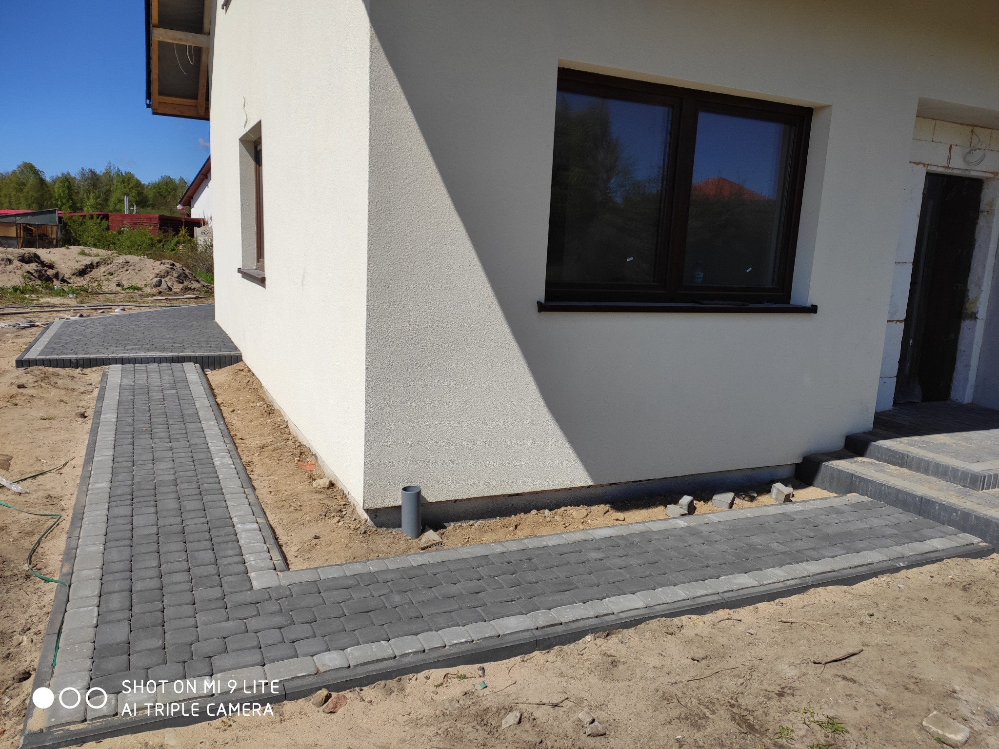 Ułożona ścieżka z szarej kostki brukowej obramowana jaśniejszą kostką, prowadząca wzdłuż białej ściany budynku z widocznymi oknami i schodami wejściowymi, w otoczeniu piasku i ziemi.