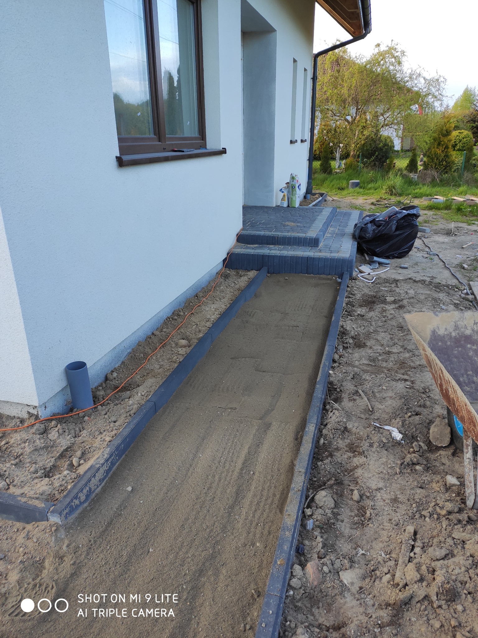 Przygotowanie podłoża pod chodnik z obrzeżami z kostki, widoczny piasek, część domu z oknem, taczka i gruz w tle.