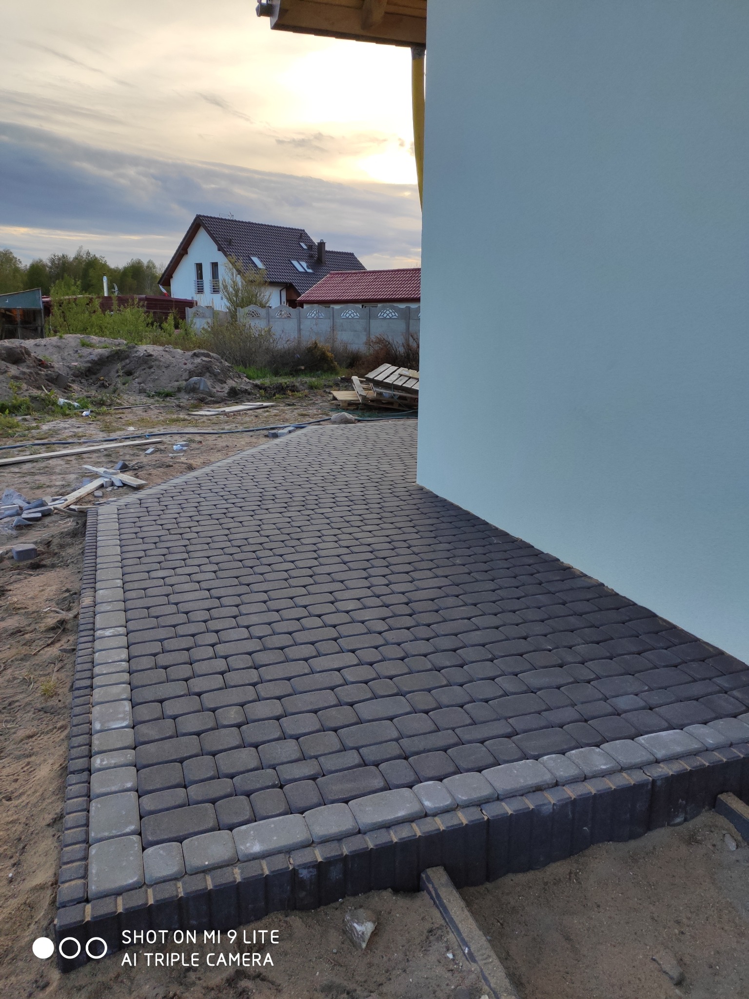 Ułożona kostka brukowa w odcieniach szarości tworząca ścieżkę przy ścianie domu, widoczne elementy wykończeniowe i piasek, w tle dom jednorodzinny i zachmurzone niebo.