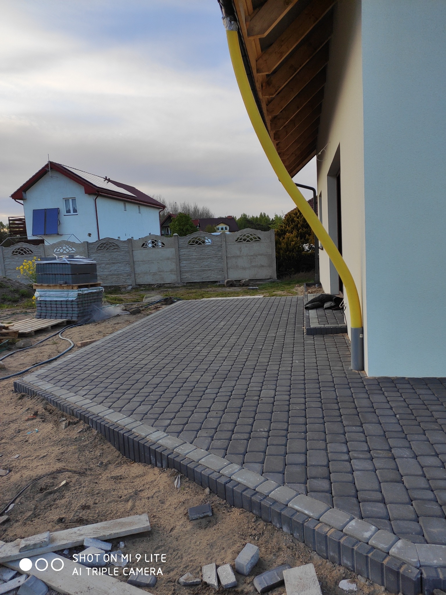 Układanie kostki brukowej na posesji, widoczny fragment nowego chodnika z kostki, obrzeża, piasek, w tle dom z panelami słonecznymi i ogrodzenie.