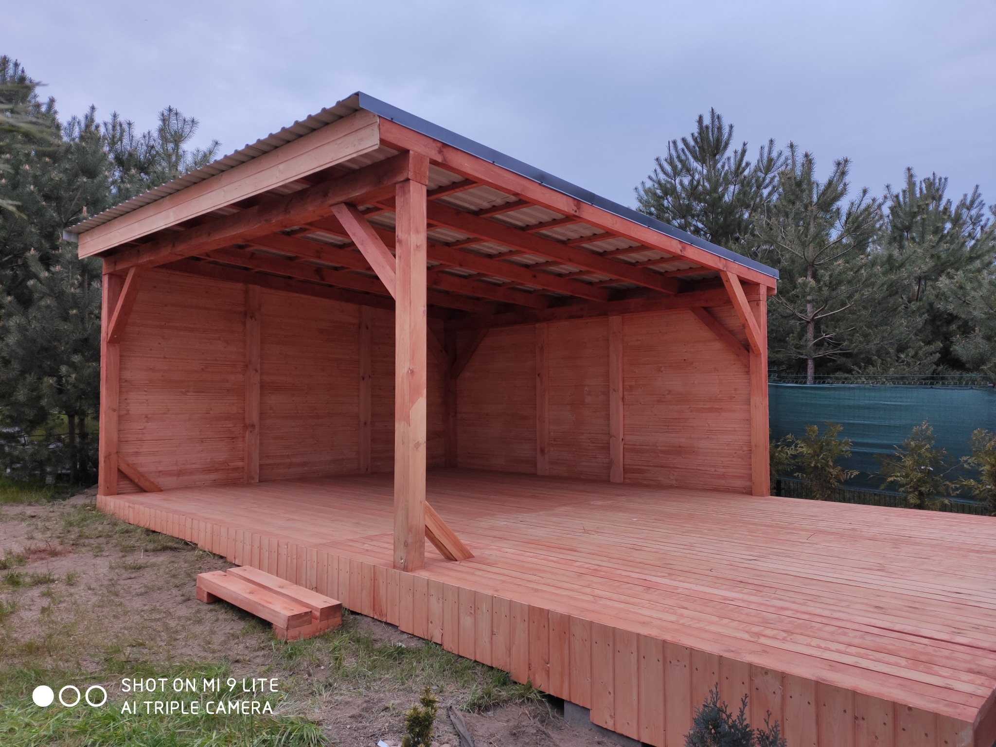 Nowa, drewniana altana z metalowym dachem, widok z przodu, na tle zieleni ogrodu i pochmurnego nieba.