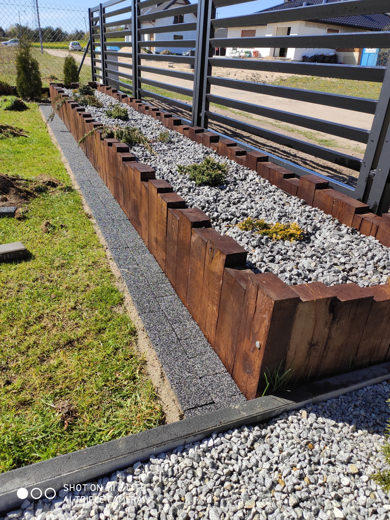 Długa rabata kwiatowa z drewnianą obudową, wypełniona szarym żwirem i niskimi roślinami iglastymi, przylegająca do czarnego ogrodzenia panelowego.