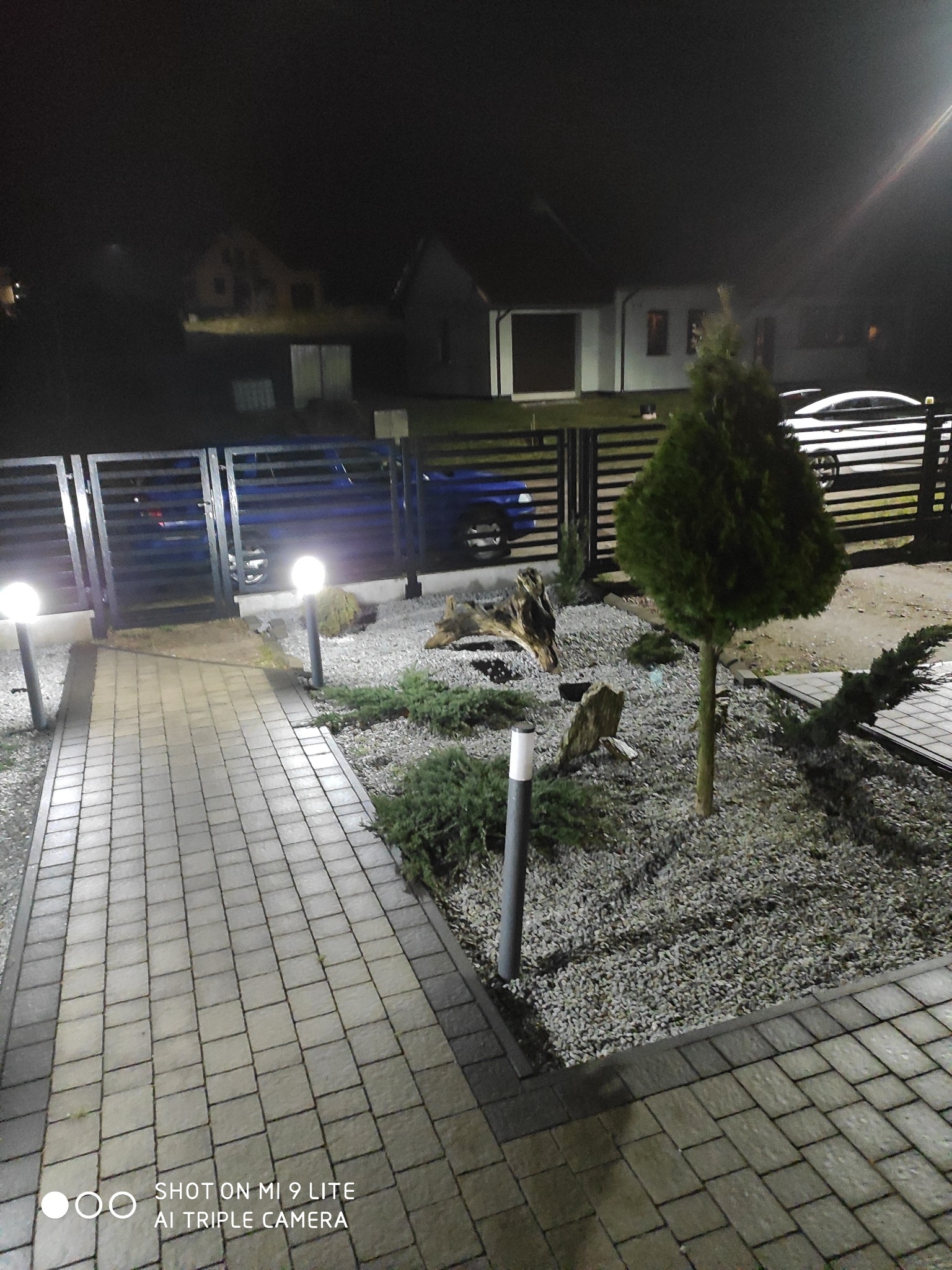 Oświetlony nocą ogród w Słupsku z ścieżką z kostki brukowej, ozdobnym żwirem, niskimi krzewami, młodym drzewkiem i elementami drewna, ogrodzony metalowym płotem.