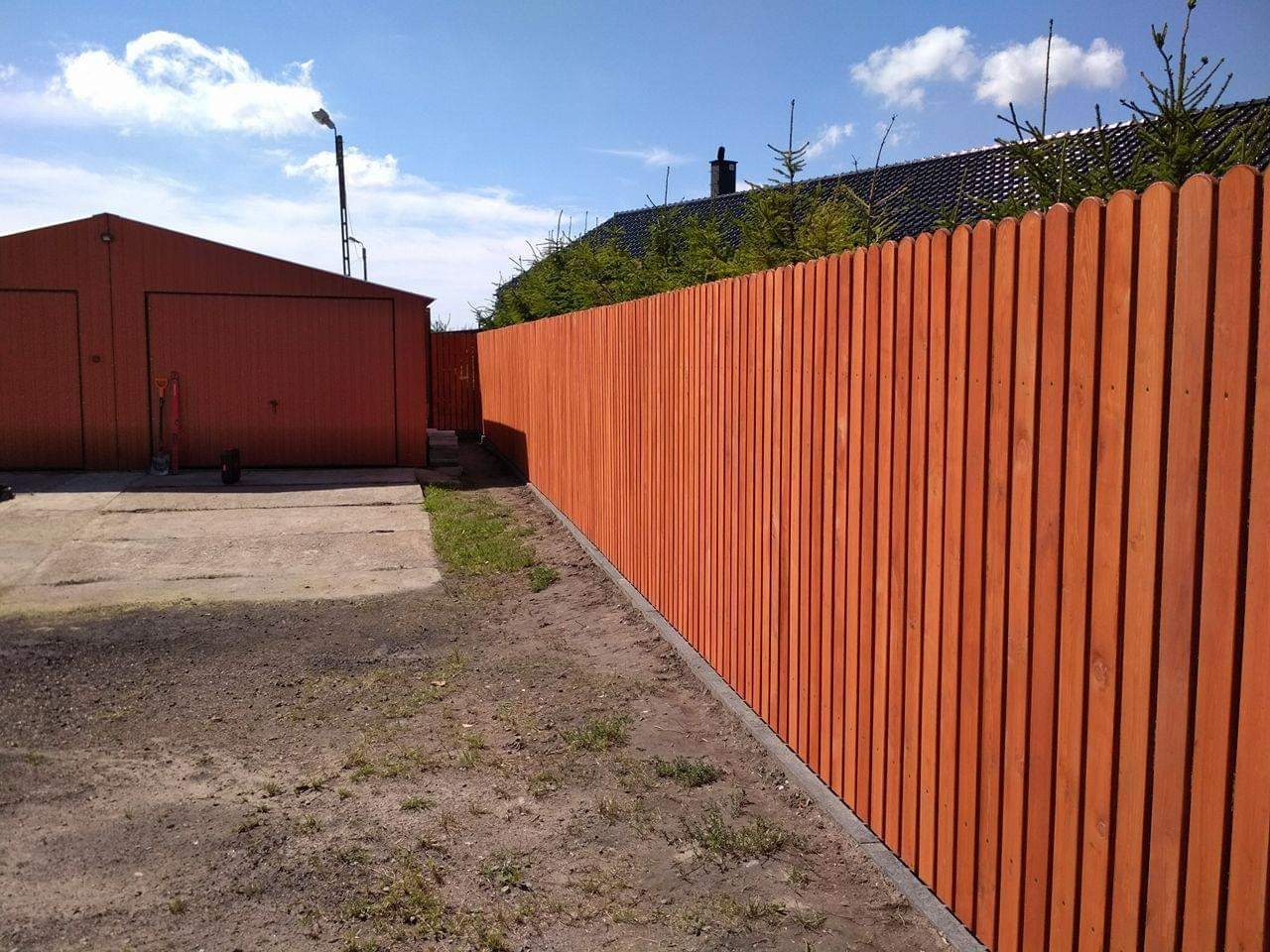 Nowy, pomarańczowy płot z desek sztachetowych, oddzielający posesję z garażem blaszanym, widoczny z perspektywy ziemi w słoneczny dzień.