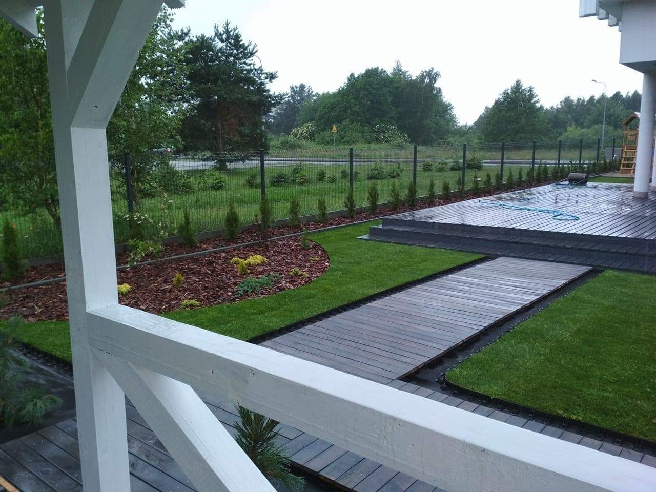 Nowoczesny ogród z drewnianym tarasem i ścieżką, świeżo posadzona trawa, rabata z korą i niskimi iglakami, widok z zadaszonego tarasu z białą balustradą