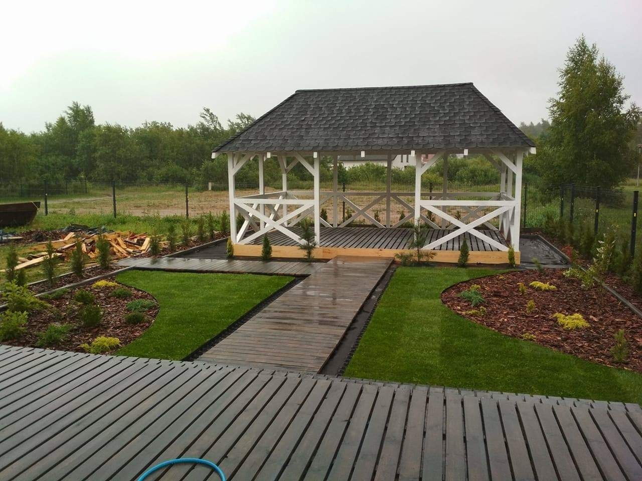 Biała altana z ciemnym dachem, otoczona świeżo założonym trawnikiem, drewnianymi ścieżkami i nasadzeniami z tujek, widok z tarasu.