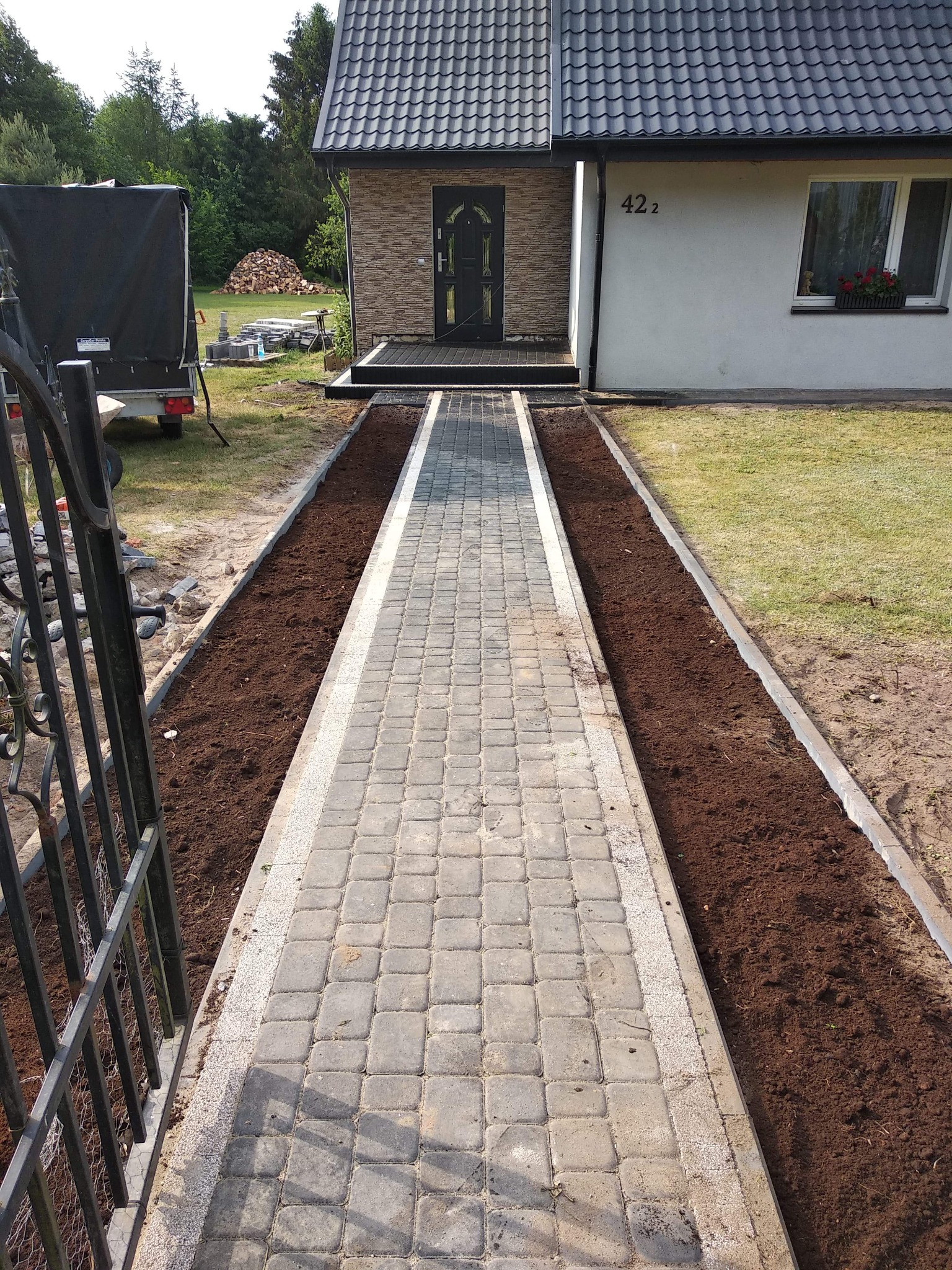 Nowo wyłożona kostka brukowa tworząca ścieżkę prowadzącą do domu z numerem 422, z przygotowanymi rabatami po bokach, widok zza metalowej bramy.