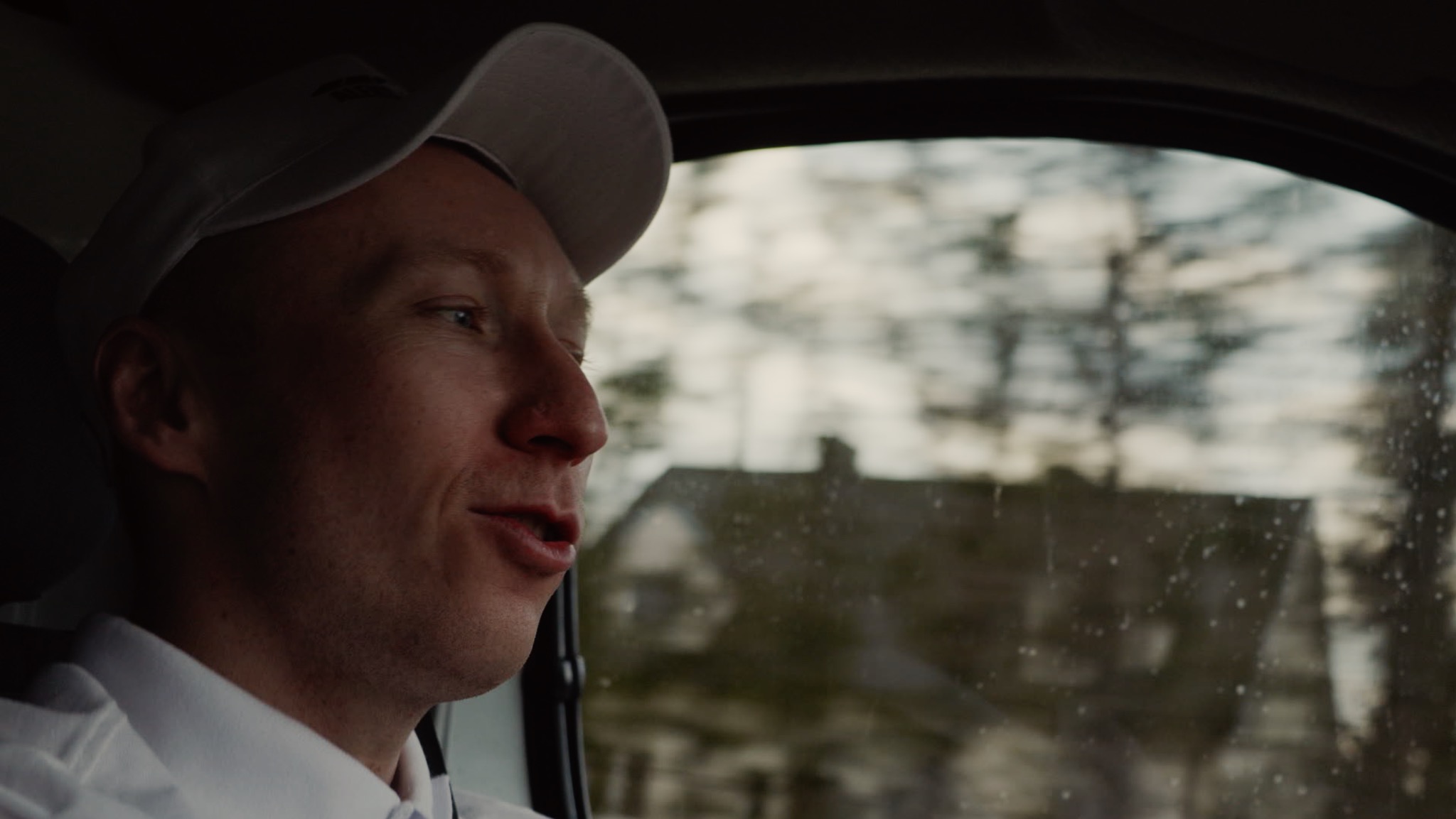 Mężczyzna w czapce i koszuli, widoczny z profilu w samochodzie, rozmawia, w tle rozmazany krajobraz z domem.