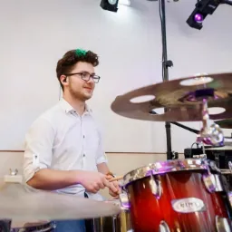 Perkusista z zielonymi pasemkami we włosach gra na perkusji marki Mapex podczas występu na scenie oświetlonej reflektorami.