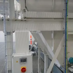 Szara metalowa konstrukcja wsporcza z korytkami kablowymi i przewodami elektrycznymi w kolorach niebieskim, żółto-zielonym i szarym, widoczna puszka elektryczna z czerwonym wyłącznikiem...