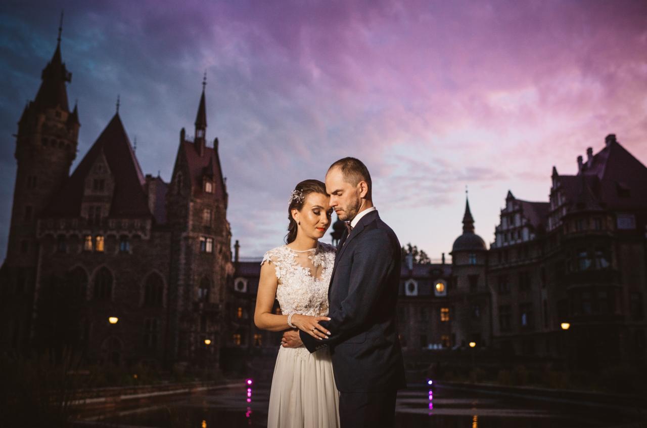 Para młoda obejmująca się na tle zamku w wieczornym świetle, panna młoda w koronkowej sukni z tiulowym dołem, pan młody w granatowym garniturze z muszką.