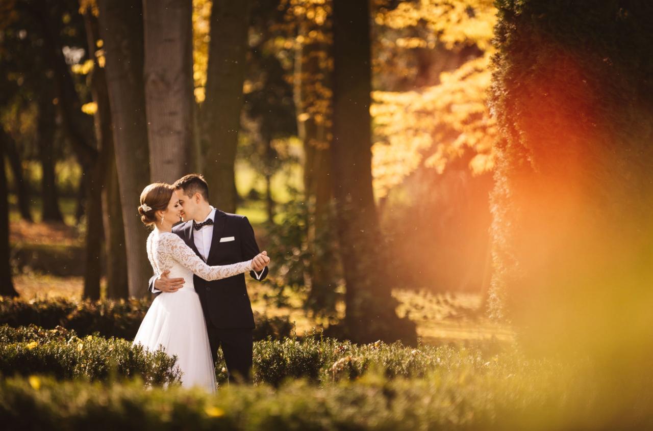 Para młoda w objęciach, pocałunek na tle parku z drzewami w jesiennych barwach, on w czarnym smokingu z muchą, ona w białej sukni z koronkowymi rękawami.